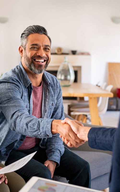 Mature indian man shaking hands with financial advisor at home. Happy smiling couple greeting broker with handshake at home. Multiethnic mid adult man and hispanic woman sealing a contract.